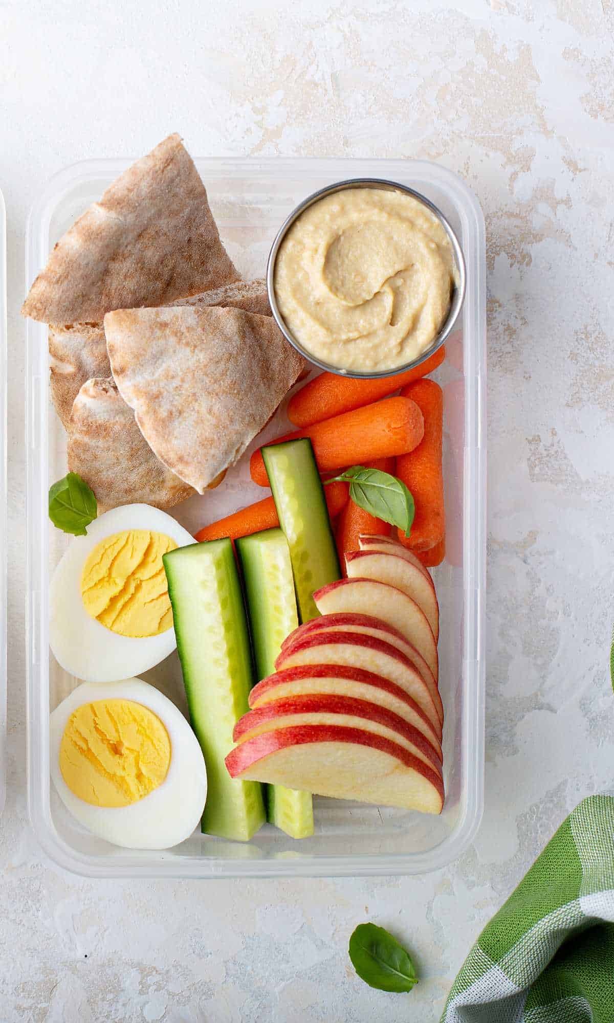 a container of healthy snacks for labor - eggs, apples, cucumbers, carrots, pita bread and hummus