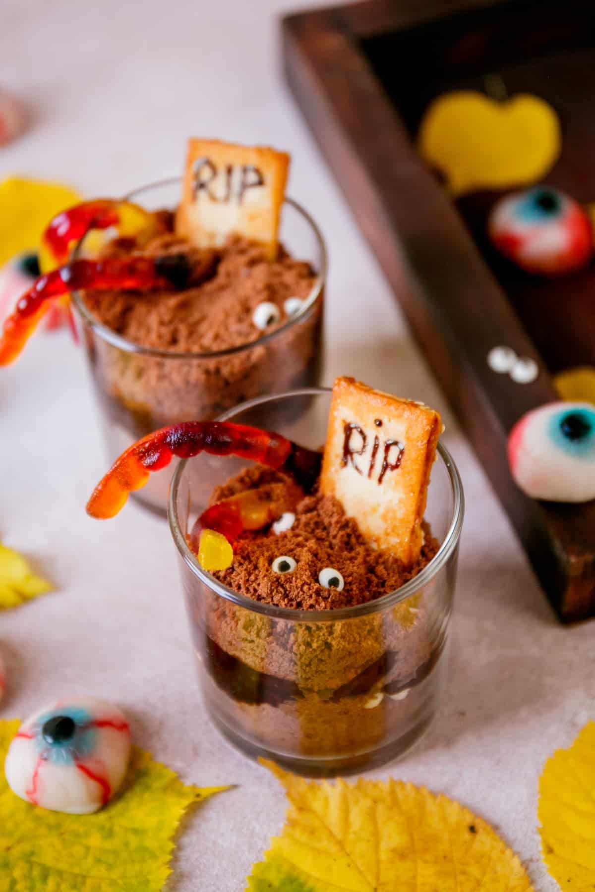 Two graveyard dirt cups with gummy worms on them and other spooky decor in the background.