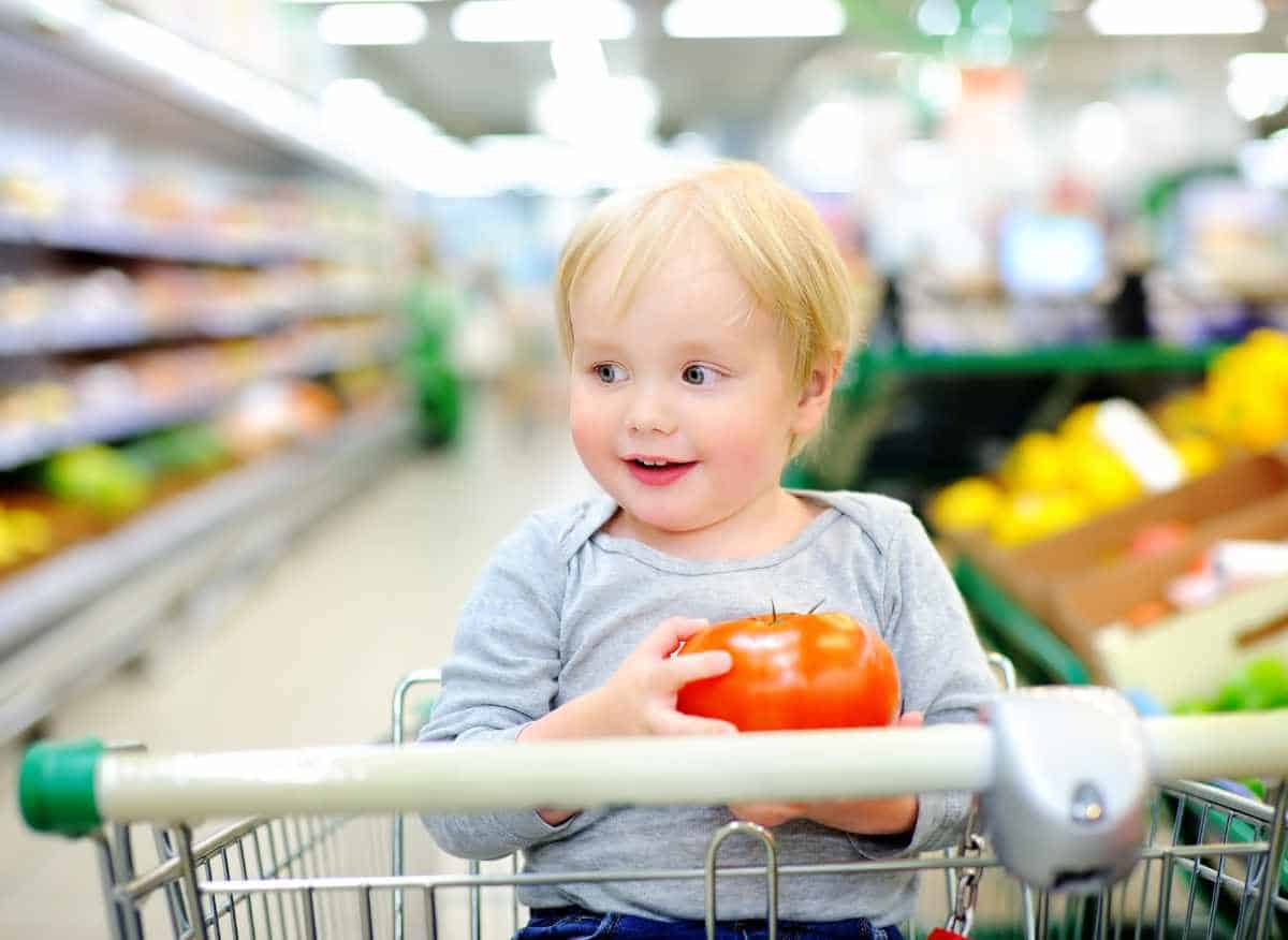 A toddler in a shopping cart holding a red tomato.