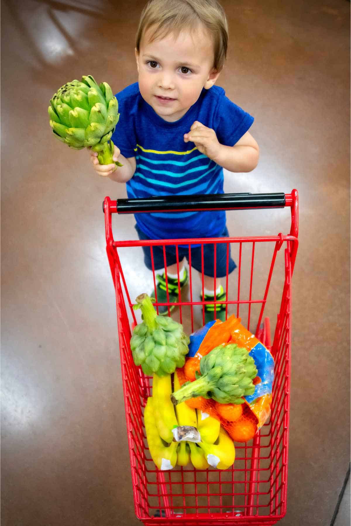 A toddler boy helps with running errands picking up vegetables and placing in his small shopping cart. 