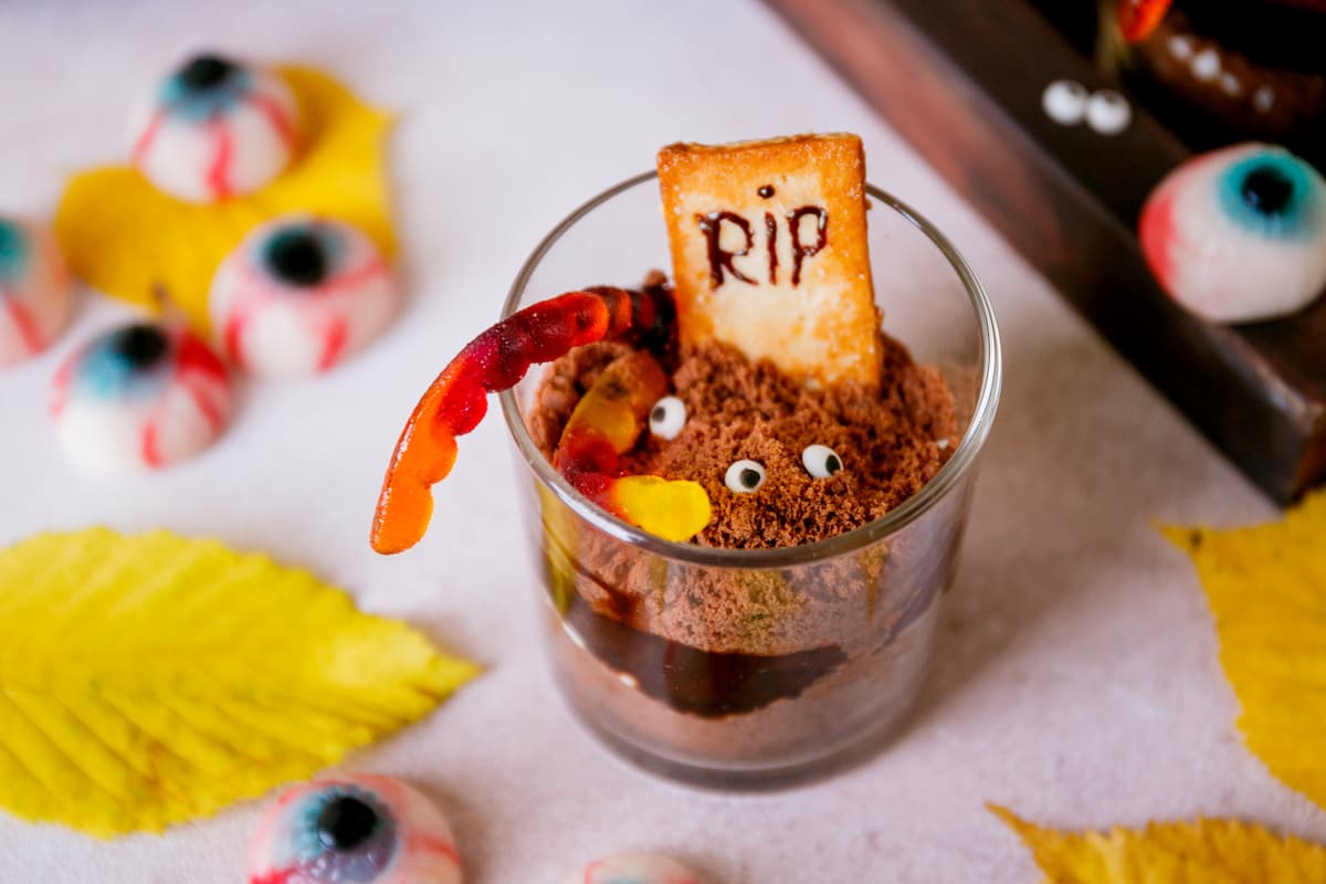 A glass of chocolate graveyard pudding with a cookie reading RIP, resembling graveyard dirt cups.