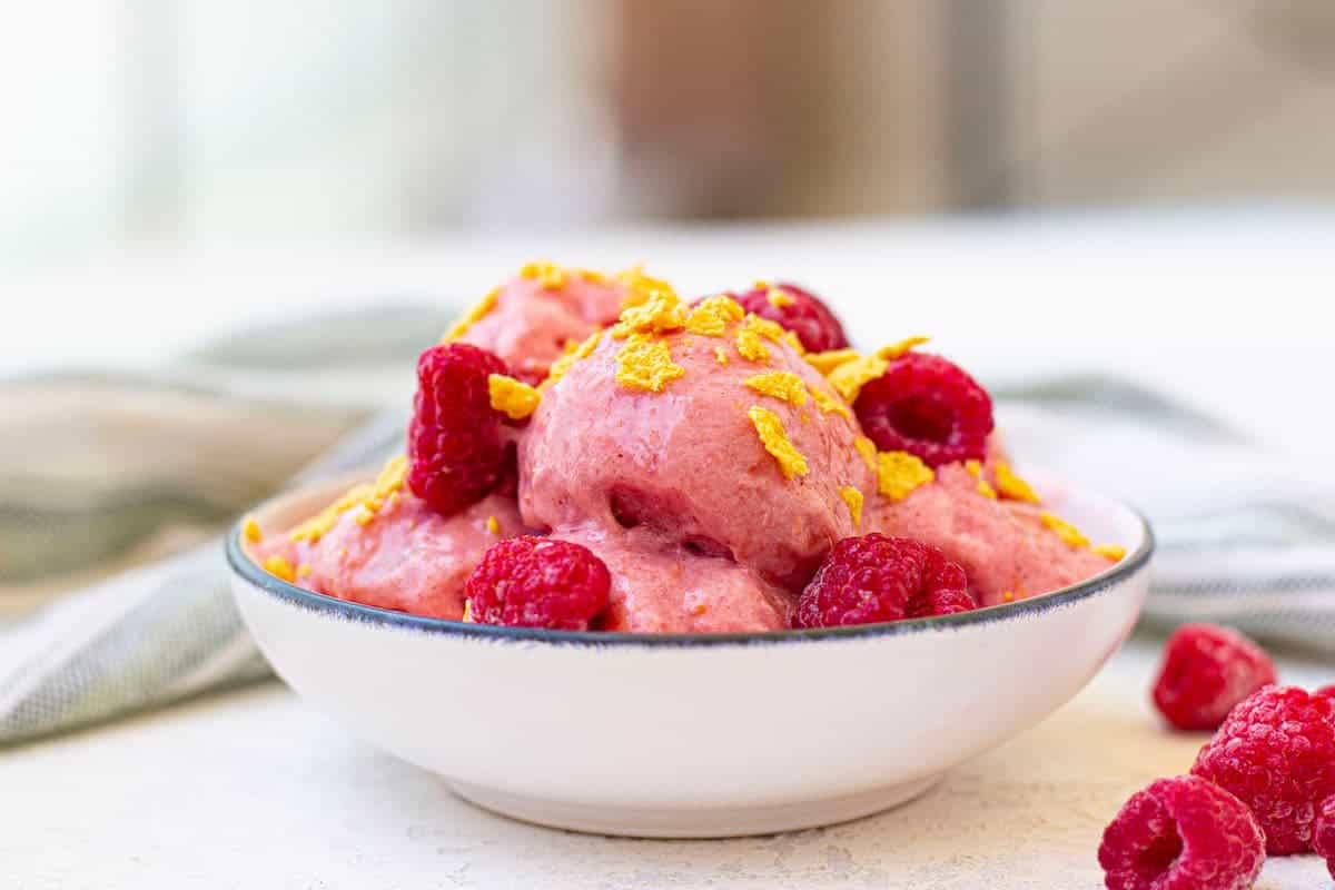 A white bowl of protein frozen yogurt garnished with raspberries and corn flakes.