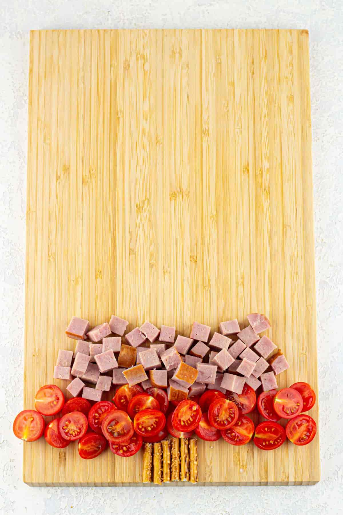 A cutting board with pretzel sesame sticks arranged as the trunk of a Christmas tree, diced beef ham, and sliced tomatoes on it.