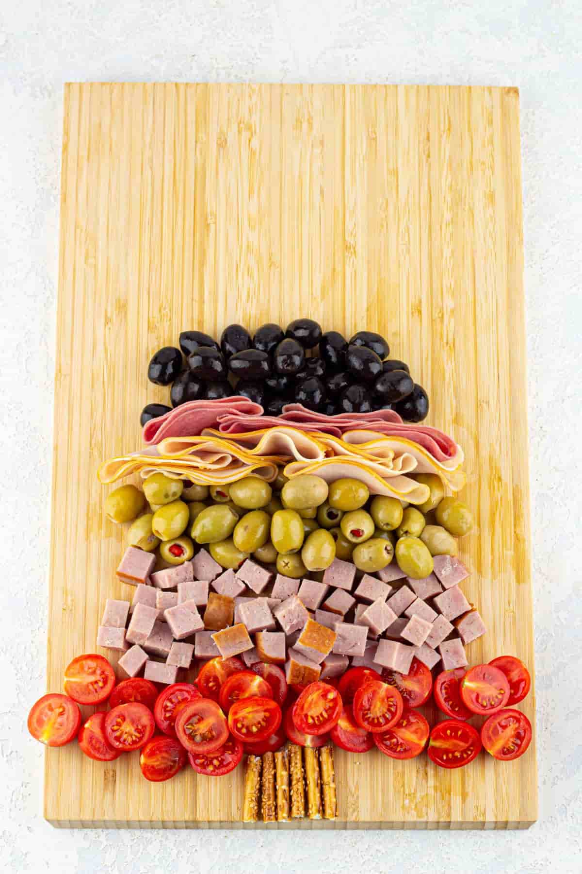 A cutting board with sesame straws, diced ham, sliced tomatoes, and green olives with chili pepper, sliced hams, and black olives on it.