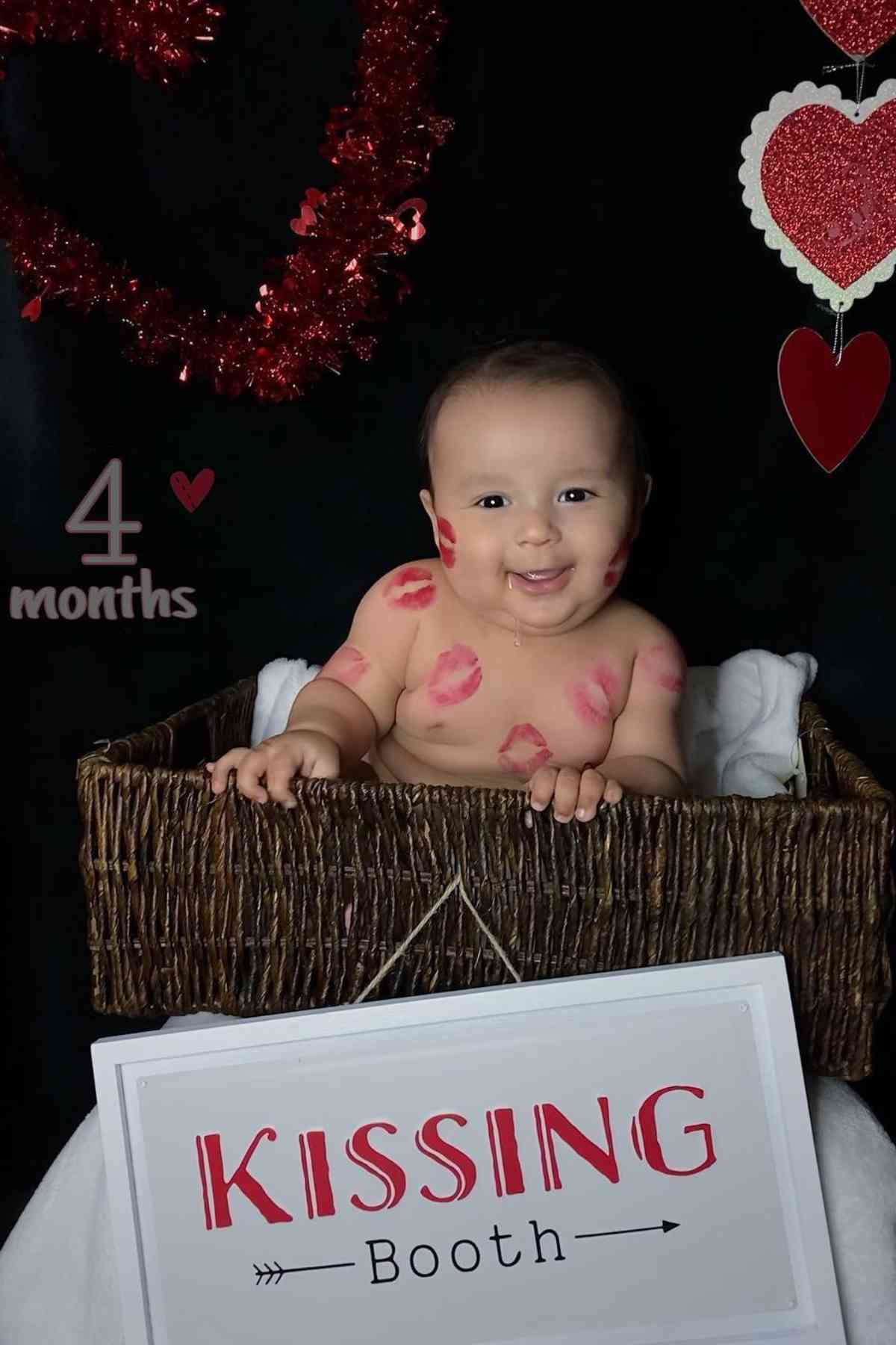 A baby sitting in a basket with a kissing booth sign.