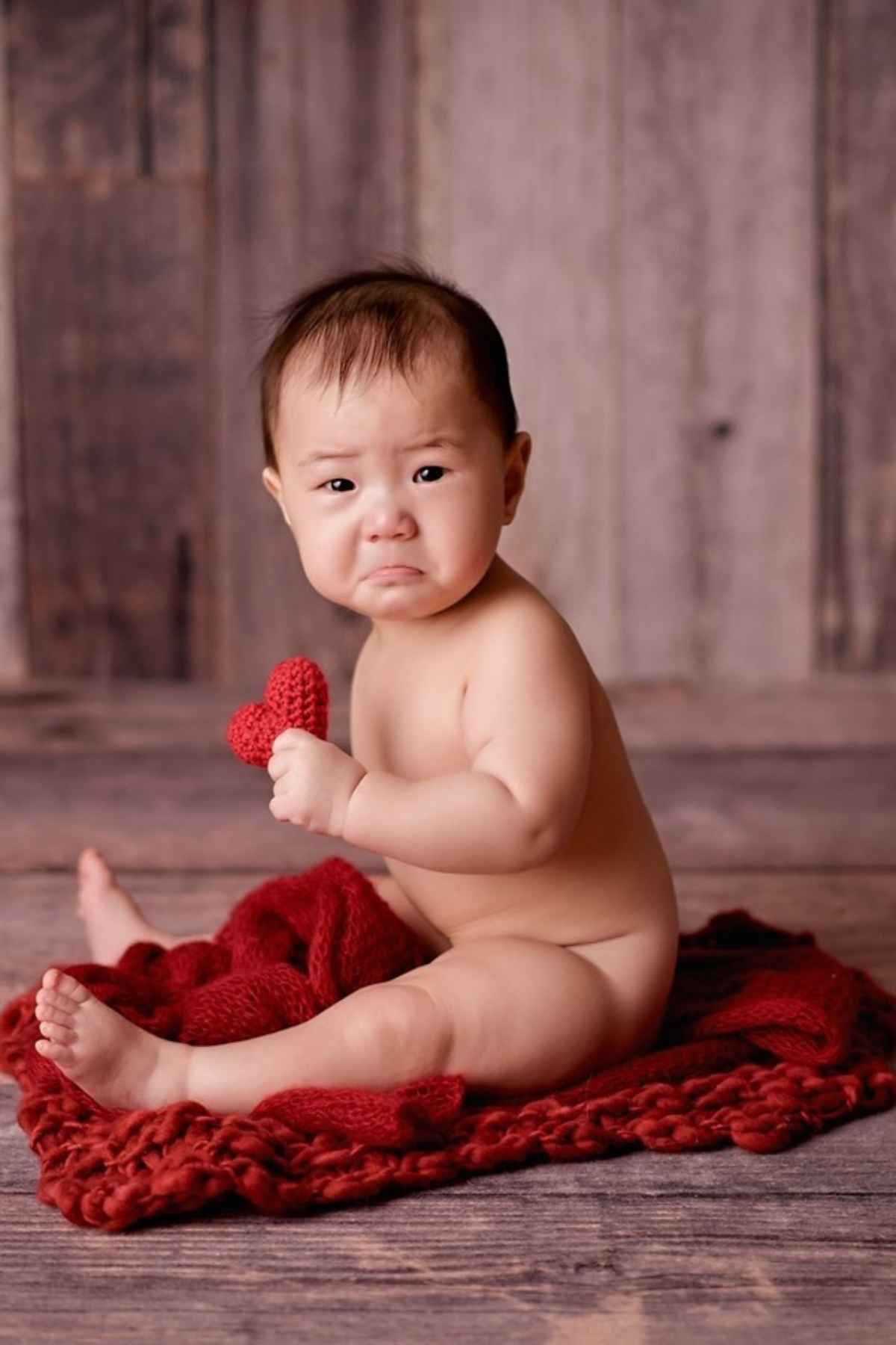 A baby sitting on a red blanket holding a heart.