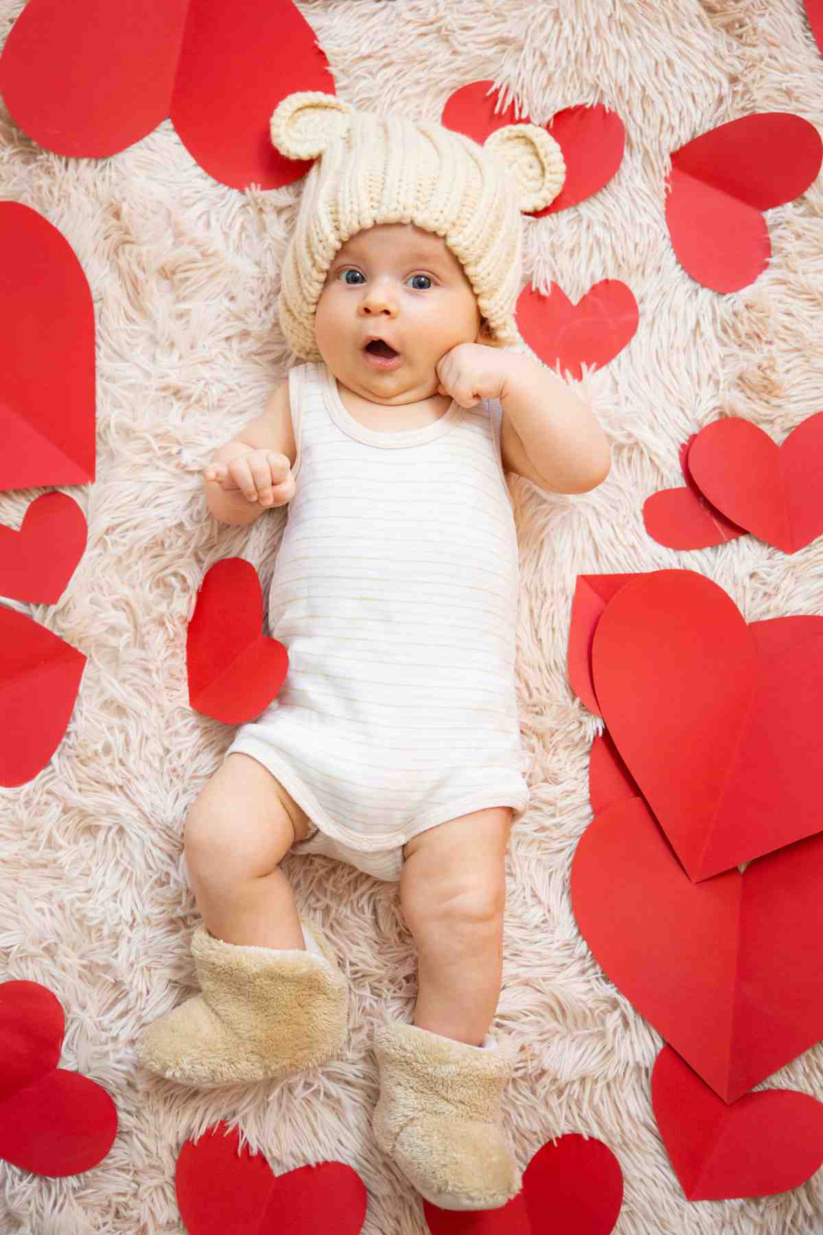 A baby wearing a teddy bear hat laying on red hearts.