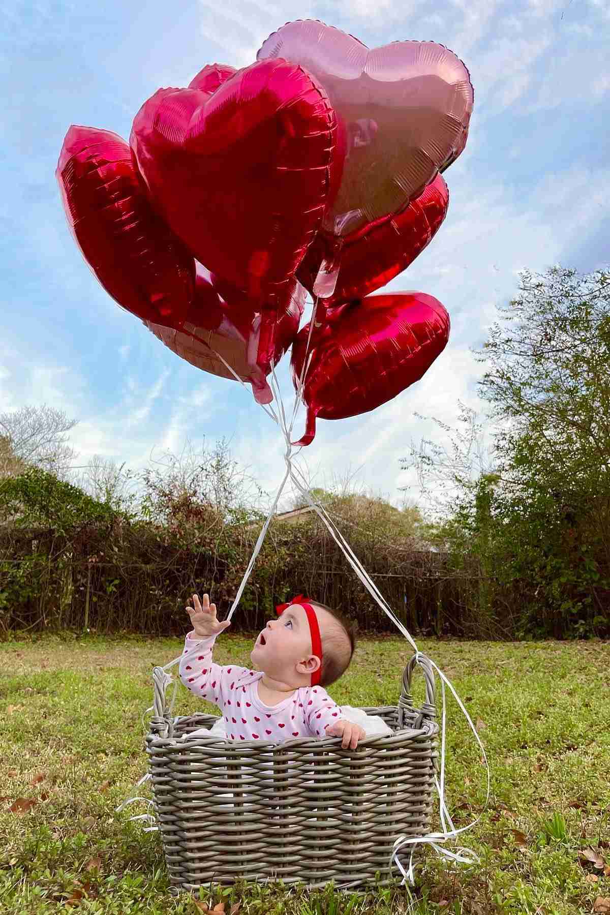 A baby laying in a basket with red heart balloons.