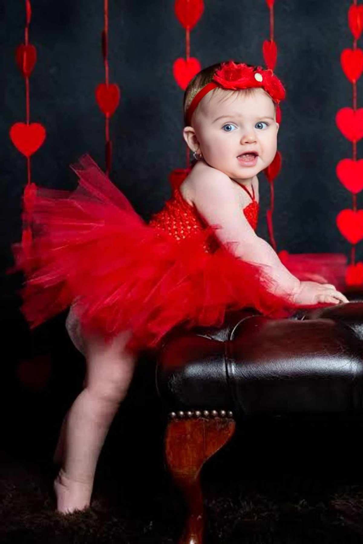 A baby girl in a red tutu standing on a leather chair.