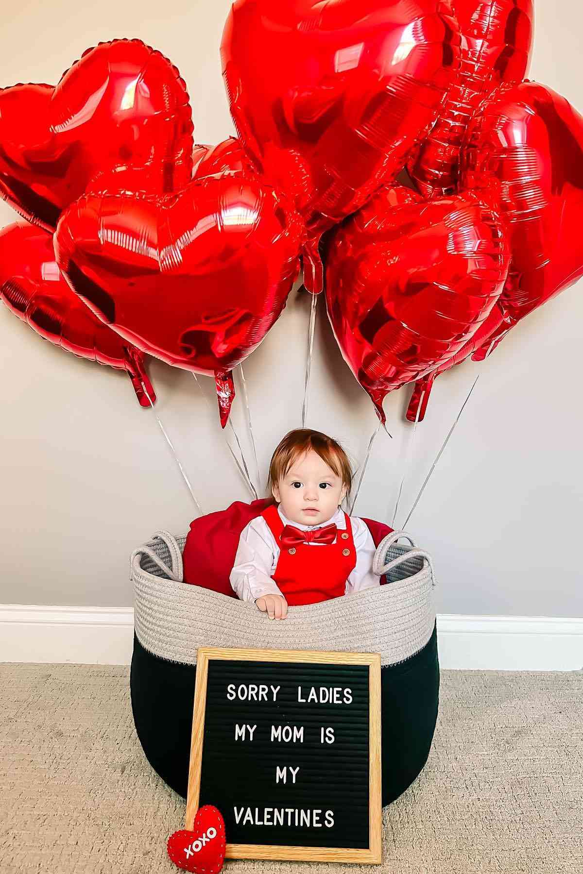 A baby sitting in a basket with heart balloons.