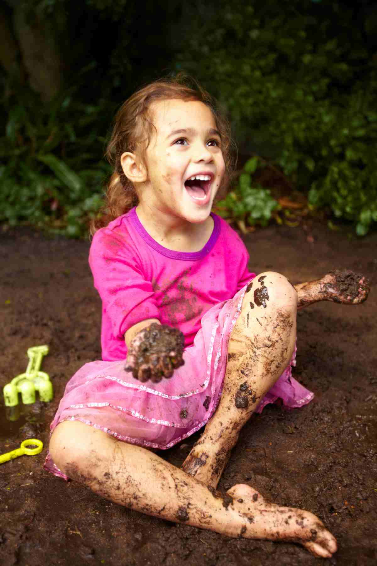 A joyful child sitting in the dirt, covered in mud, with toys nearby.