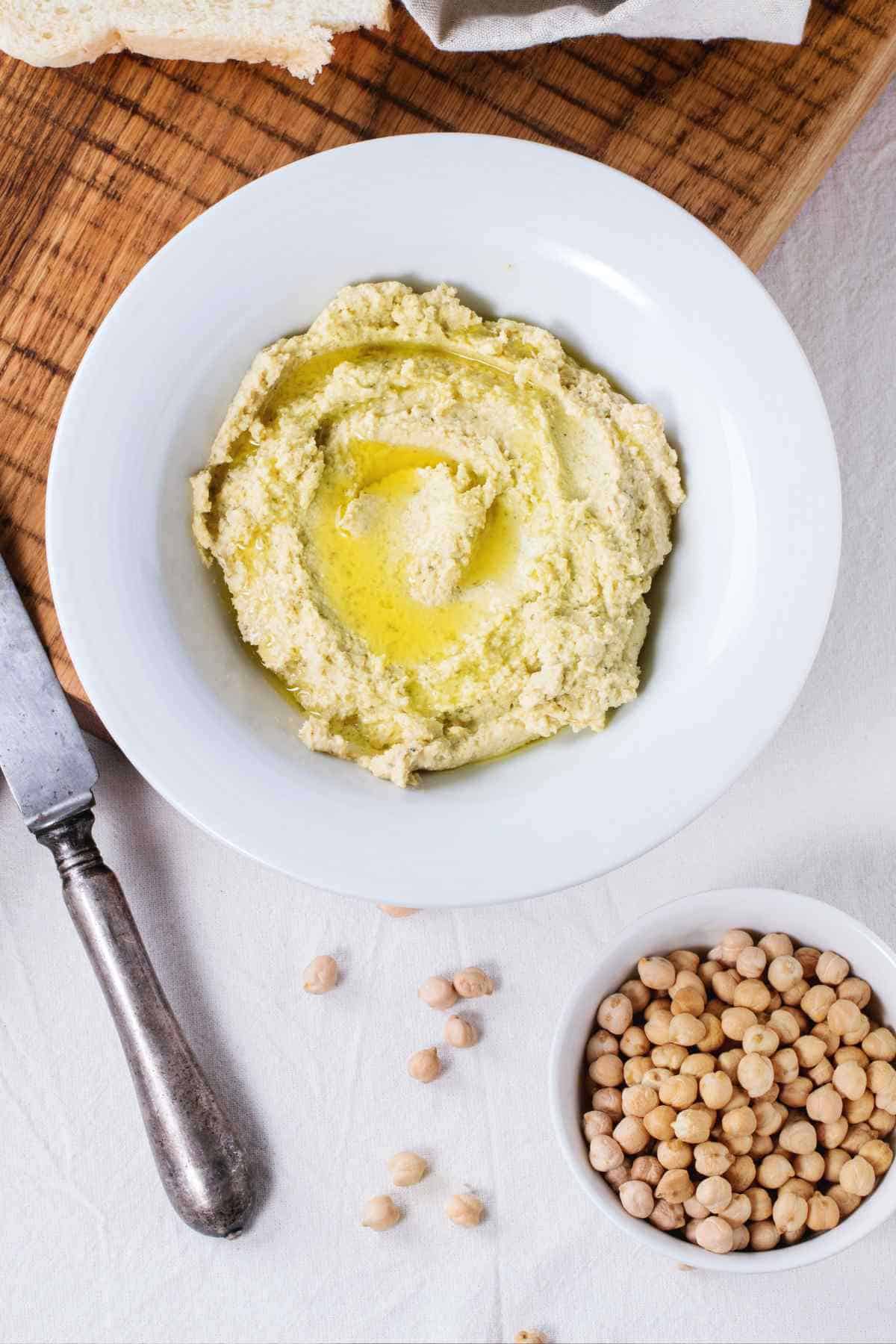 A bowl of freshly made hummus garnished with olive oil, accompanied by whole chickpeas and side.