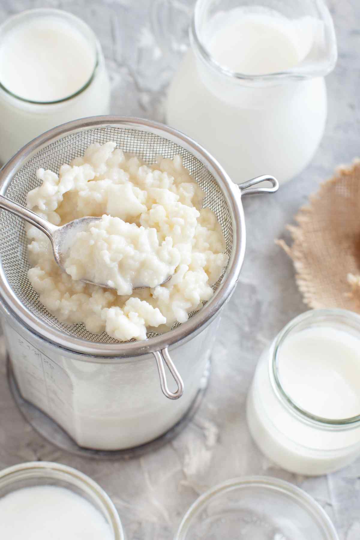 Fresh kefir grains being strained with a spoon over a jar, with bottles of milk on the side.