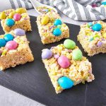 A slate tray of M&M rice krispie treats topped with pastel-colored candies and sprinkles.