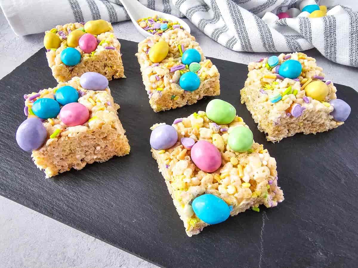 A slate tray of M&M rice krispie treats topped with pastel-colored candies and sprinkles.