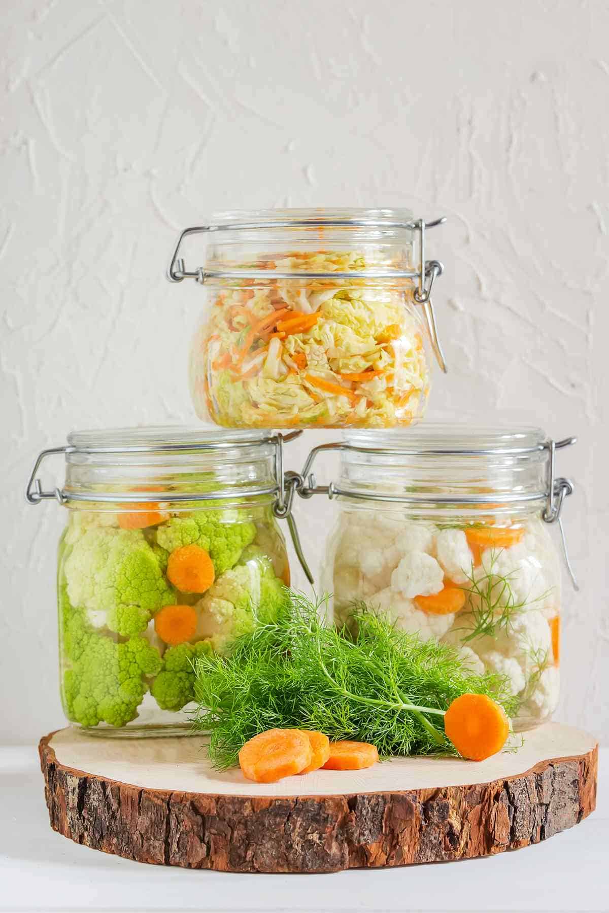 Three glass jars containing various pickled vegetables arranged on a wooden slice.