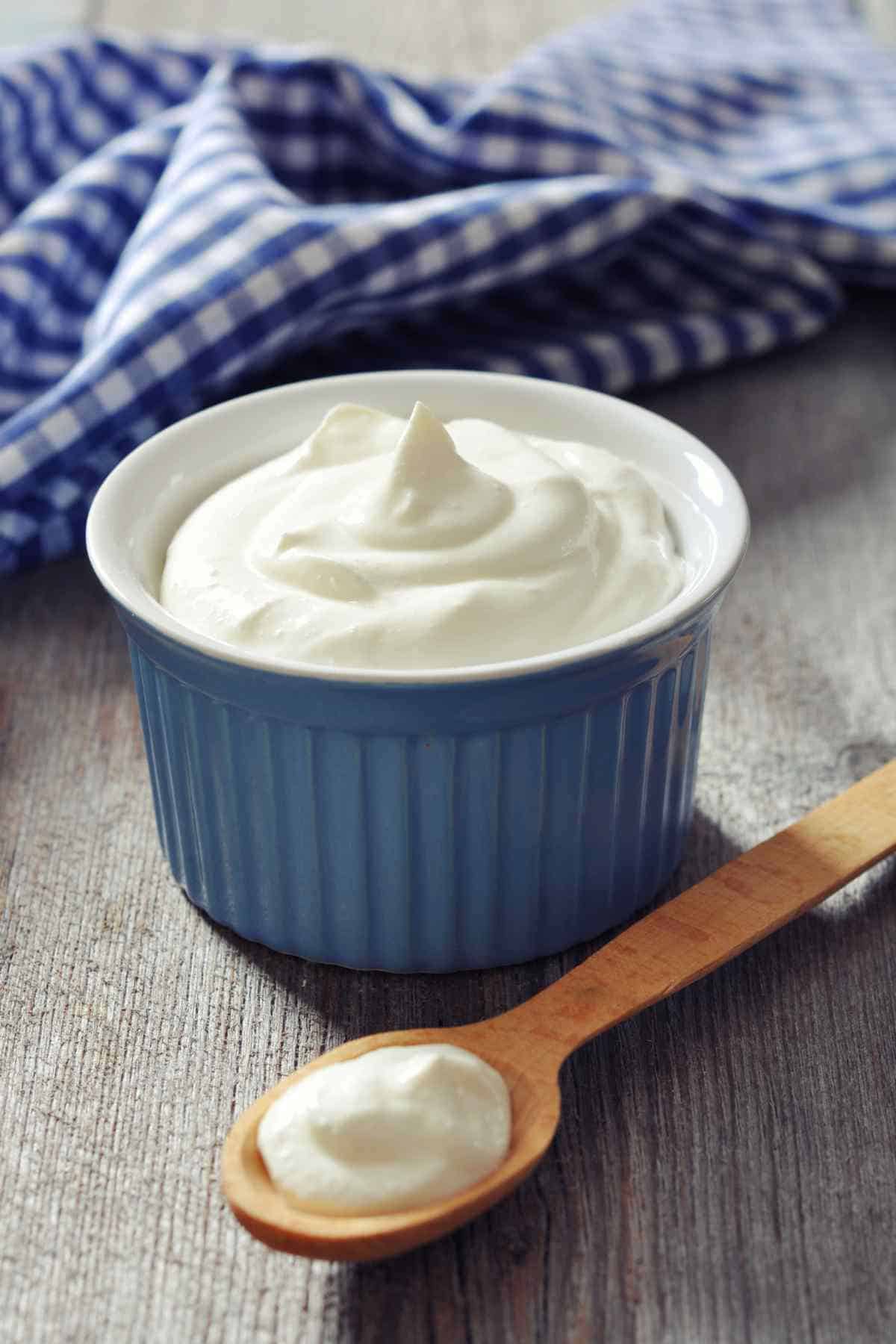 A bowl of creamy yogurt with a wooden spoon on a rustic wooden surface, accompanied by a blue and white checked napkin.
