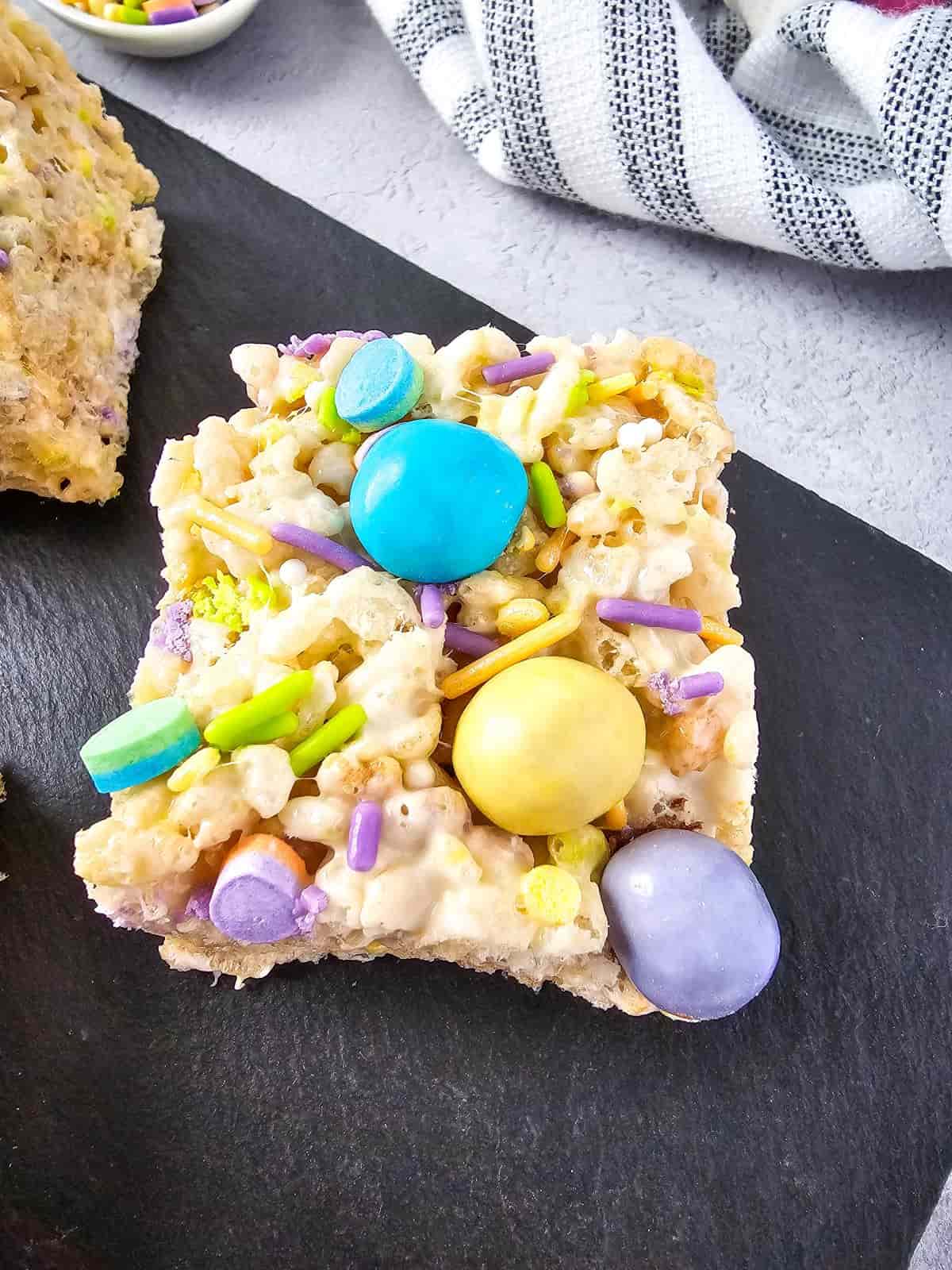 M&Ms rice krispie treat topped with colorful candy and sprinkles on a serving board.