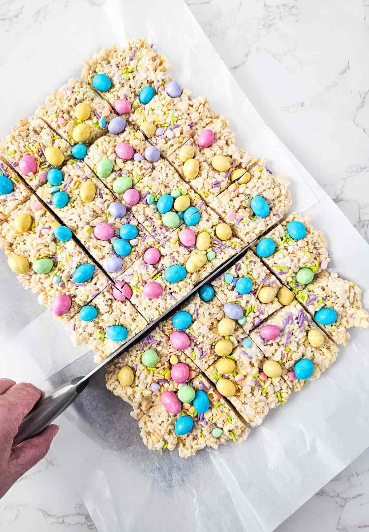 Slicing a tray of rice cereal treats decorated with pastel-colored M&M peanut candies.