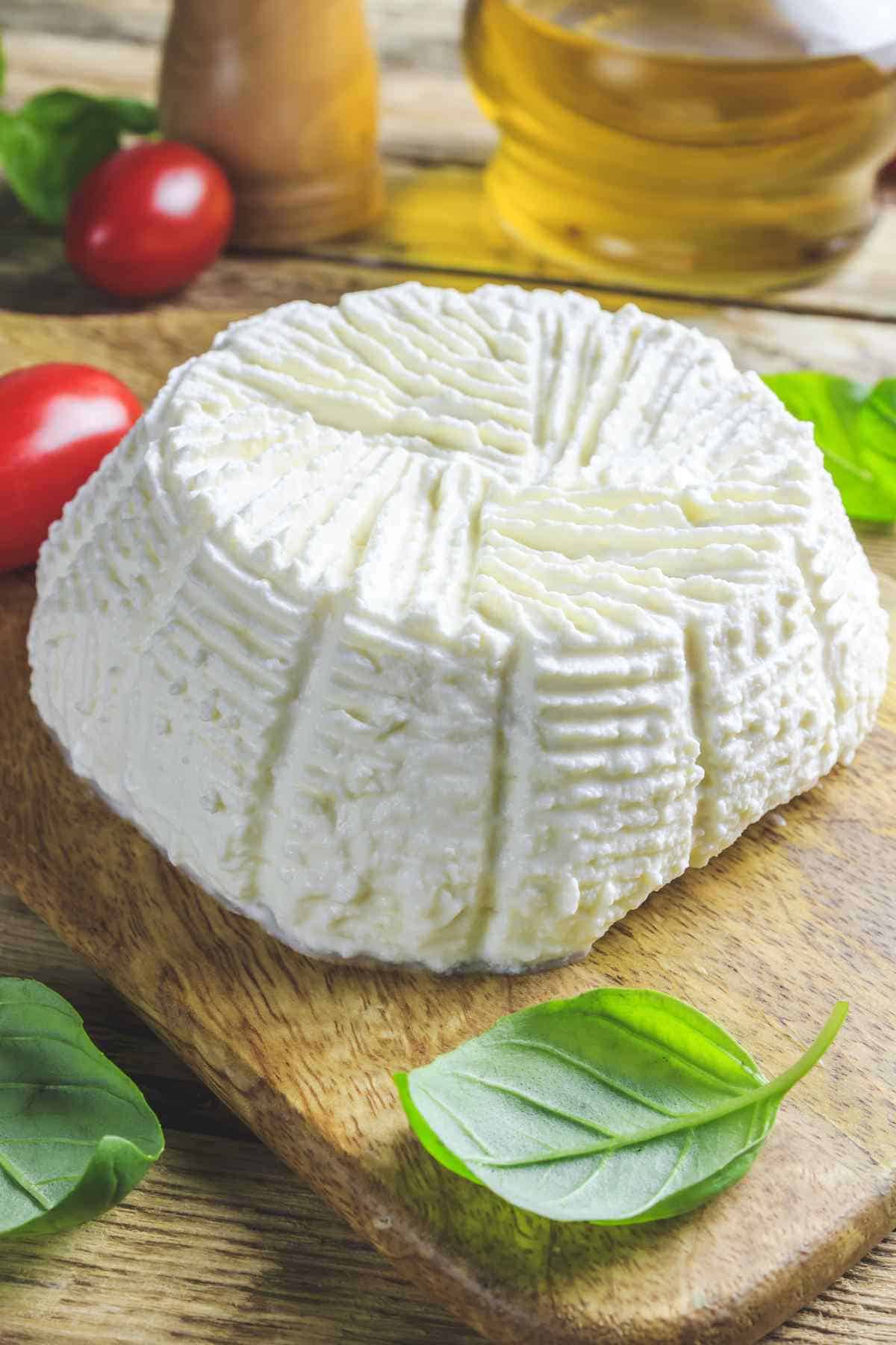 Fresh ricotta cheese on a wooden board with basil leaves and tomatoes in the background.