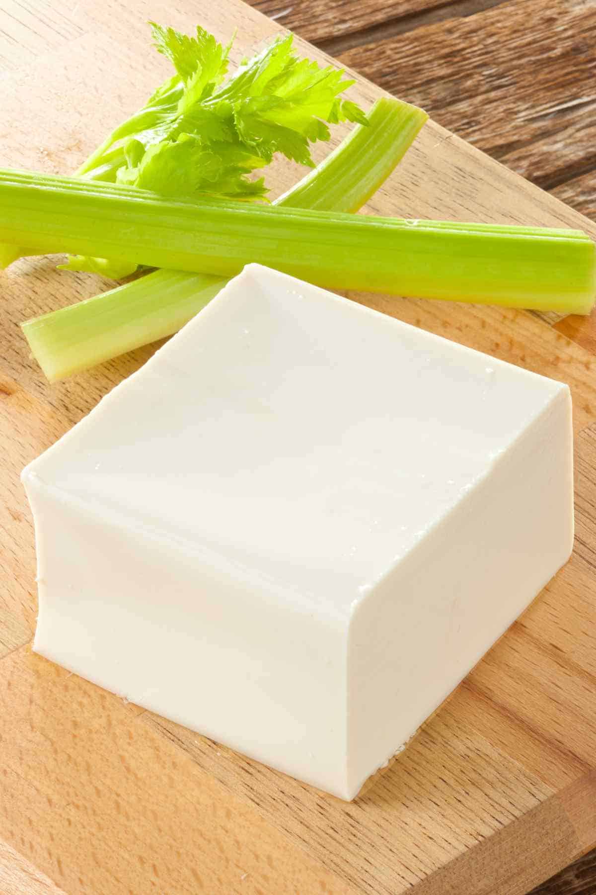 A block of tofu on a wooden cutting board accompanied by stalks of celery.