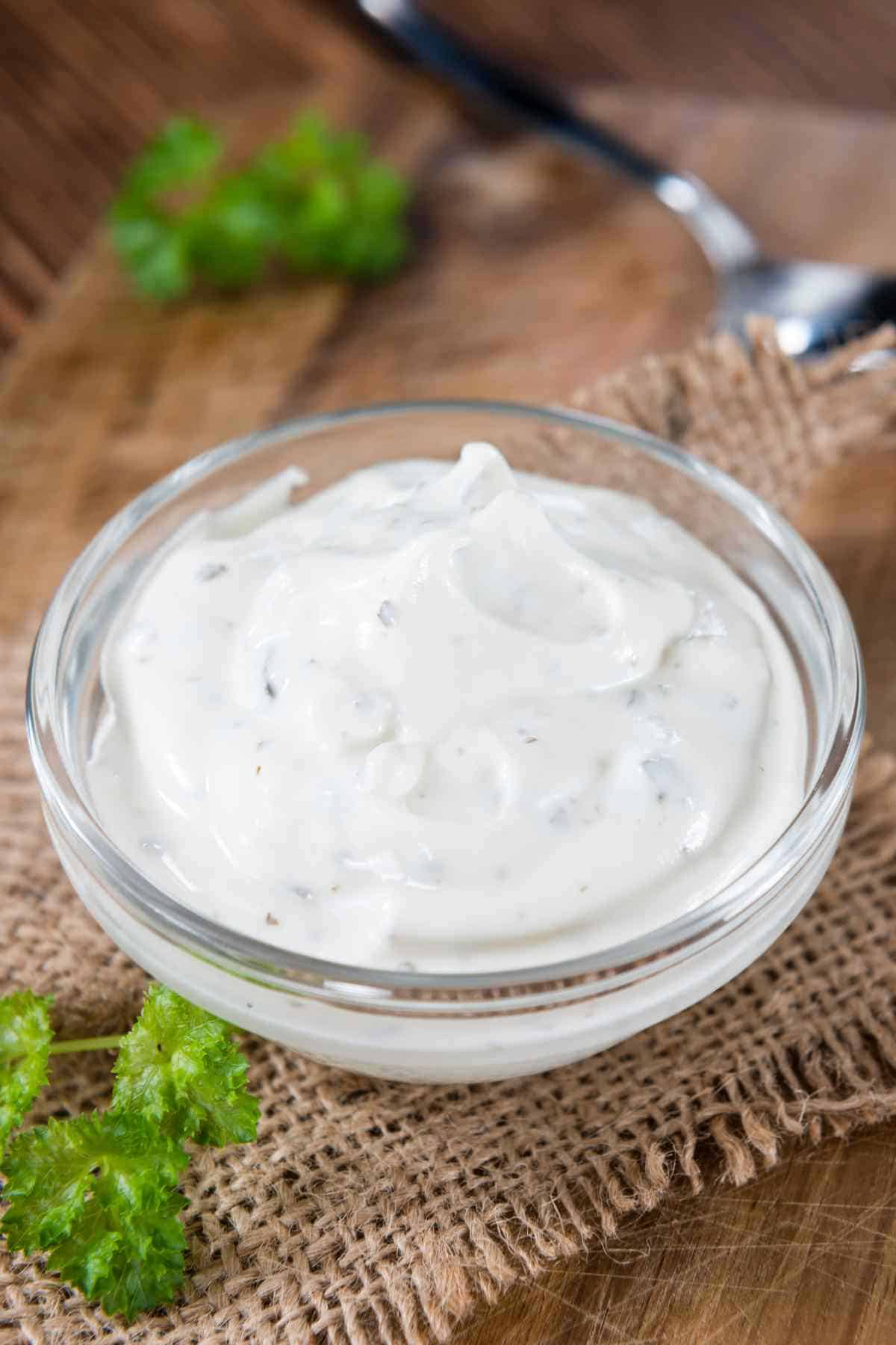 A bowl of sour cream on a wooden surface with a spoon and green garnish in the background.