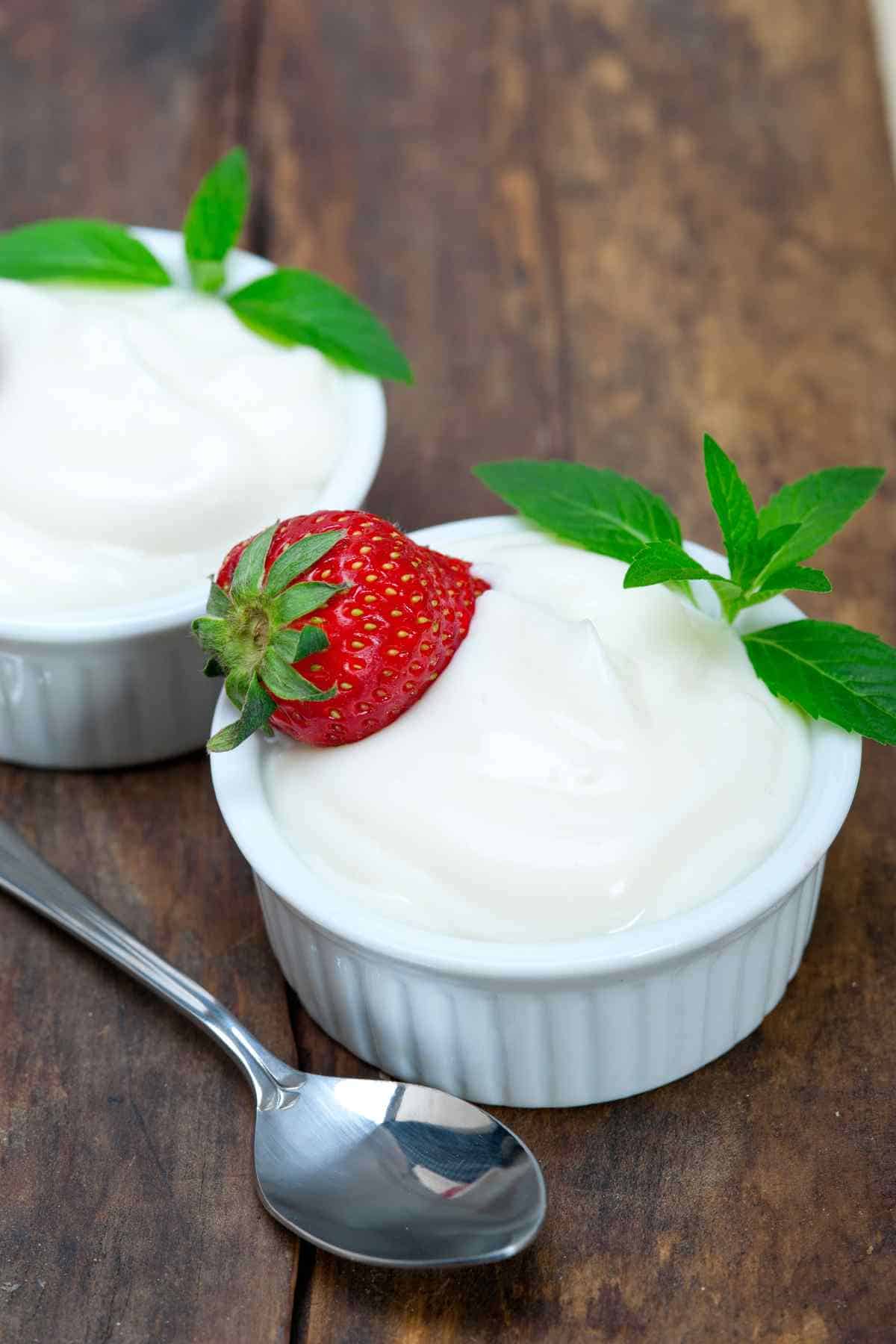 Two bowls of yogurt garnished with strawberries and mint leaves on a wooden surface with a spoon beside them.