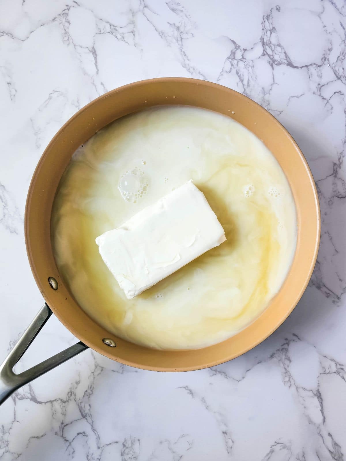 A block of cream cheese melting on a large skillet with milk and chicken broth.