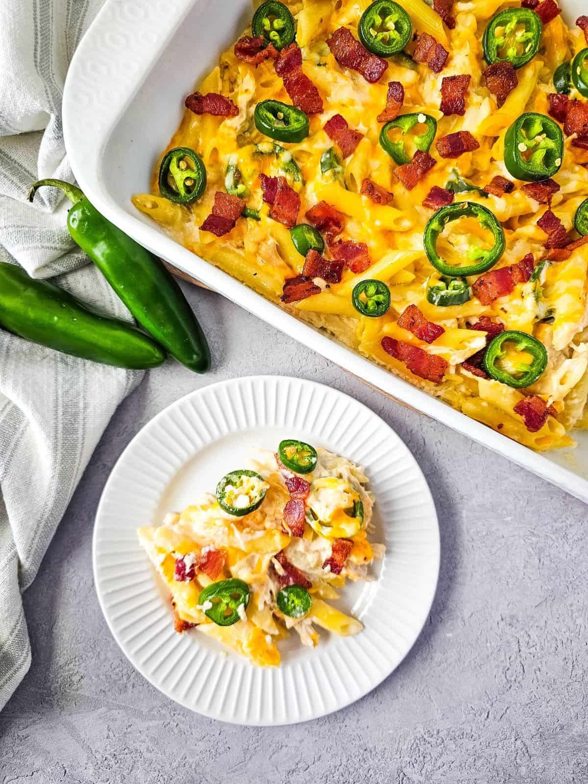 A plate of baked jalapeño poppers chicken casserole topped with bacon bits and sliced jalapeños, with the full dish in the background and fresh peppers on the side.