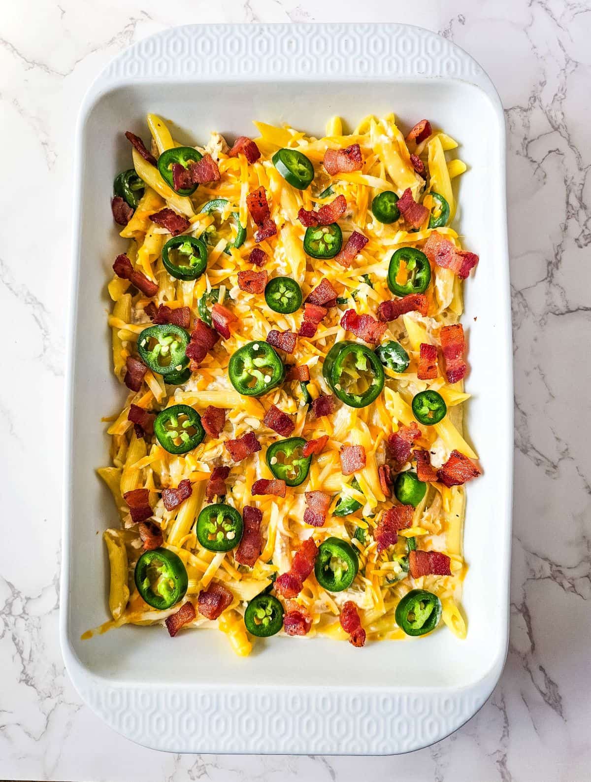 Jalapeño poppers chicken in a casserole dish with cream cheese mixture topped with shredded cheese, sliced jalapeños, and chopped bacon.