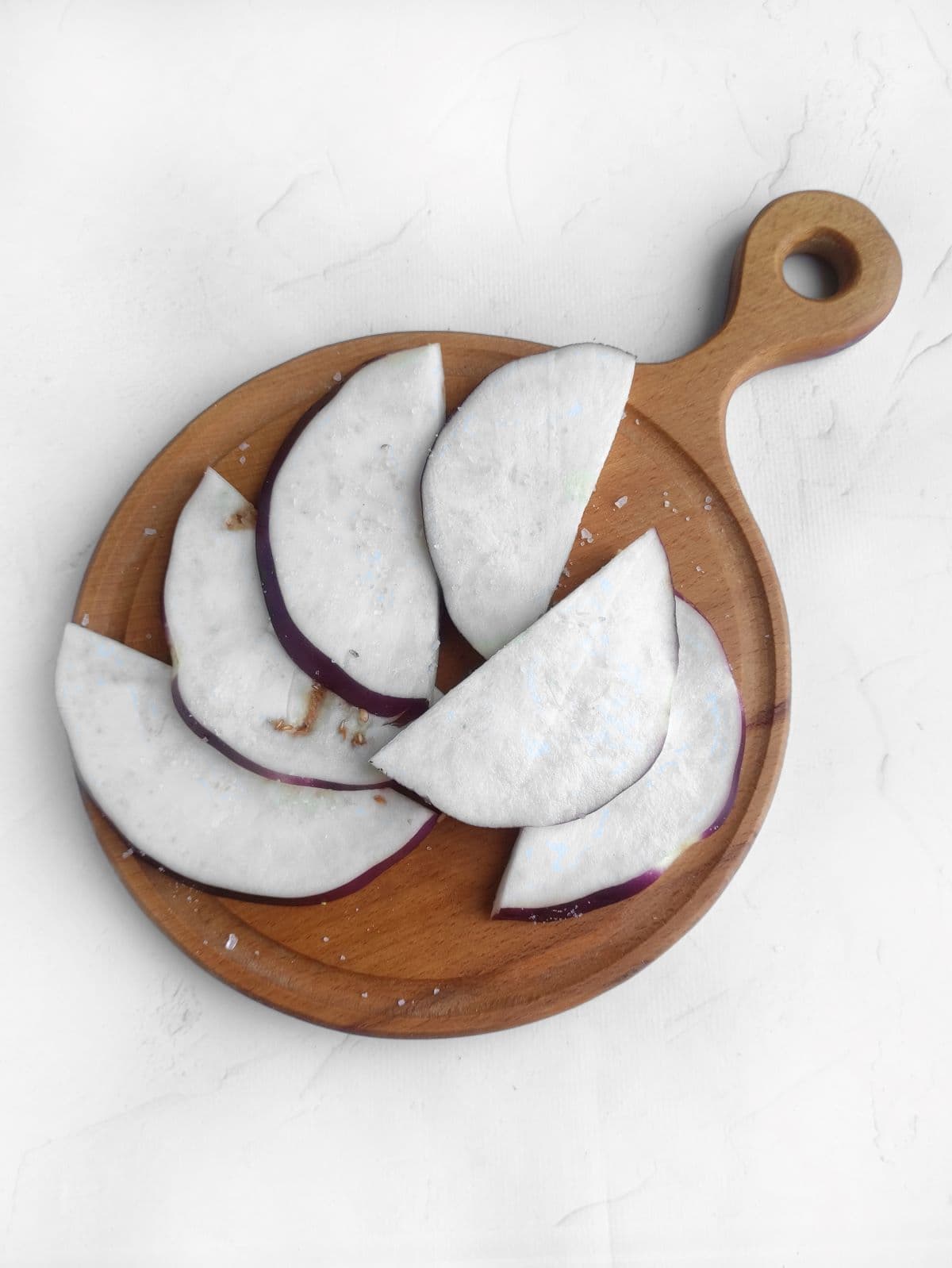 Sliced eggplant on a wooden cutting board, scattered with salt, against a white background.