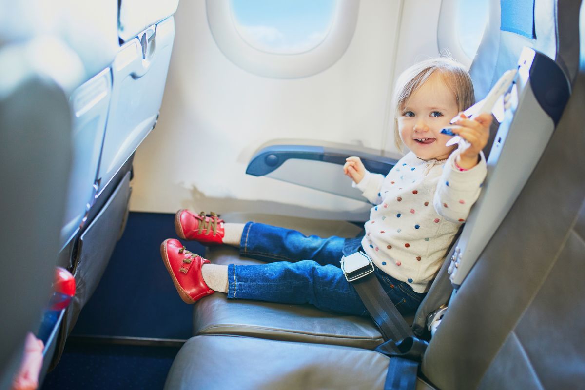 Toddler sitting in an airplane seat, wearing a polka dot shirt and red shoes while holding a toy plane.
