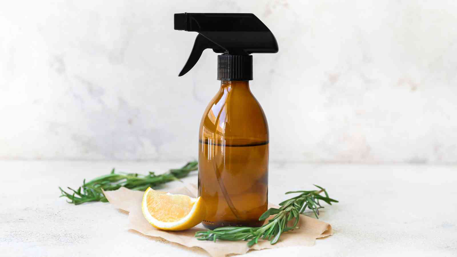 A brown glass spray bottle with a black trigger nozzle is placed on a piece of brown paper along with a lemon wedge and two sprigs of rosemary.