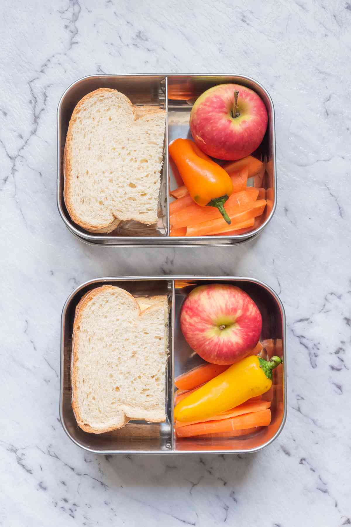Two lunch containers each with a sandwich, an apple, a small yellow pepper, and sliced carrots are perfectly placed on a marble countertop—ideal for a road trip with kids.