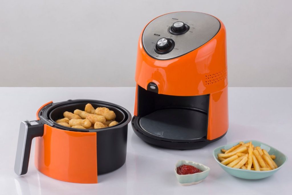 An orange air fryer with a basket of cooked nuggets, a small saucer of fries, and a small bowl of ketchup on a white surface.