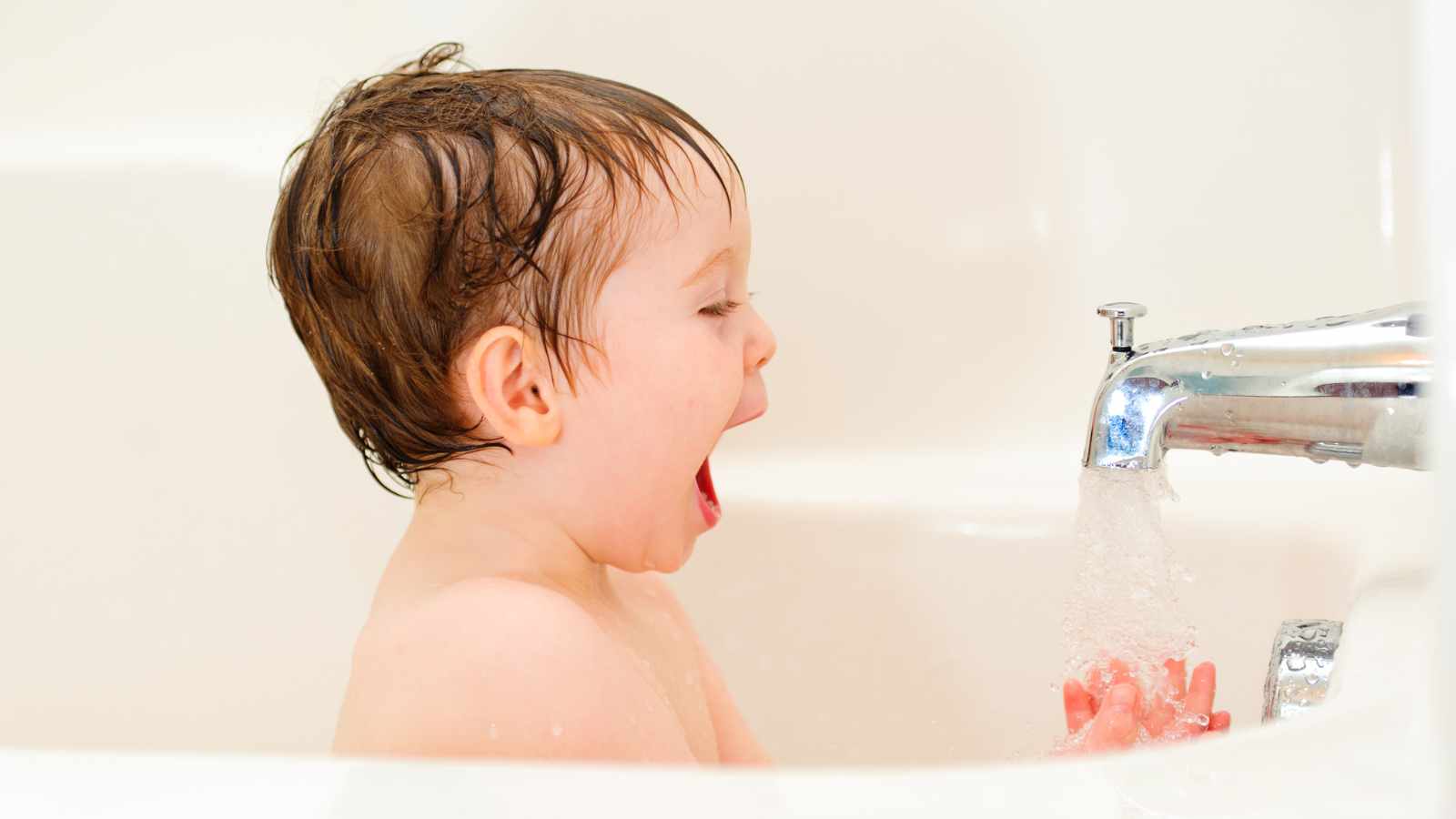 A young child, with wet hair holds their hands under running water from a bathroom faucet.
