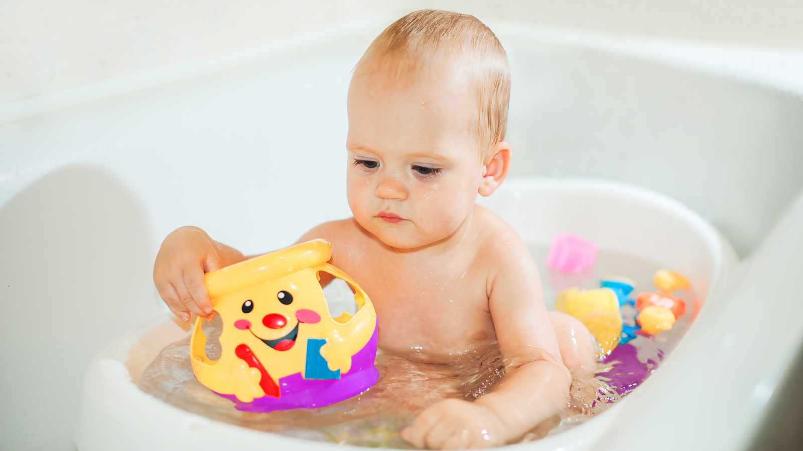 A baby sitting in a bathtub is holding a colorful toy shaped like a smiling face.