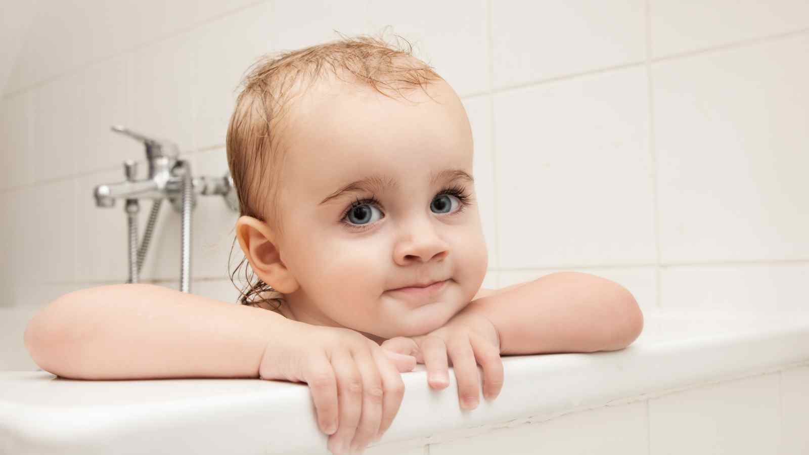A baby with wet hair rests its arms on the edge of a bathtub.