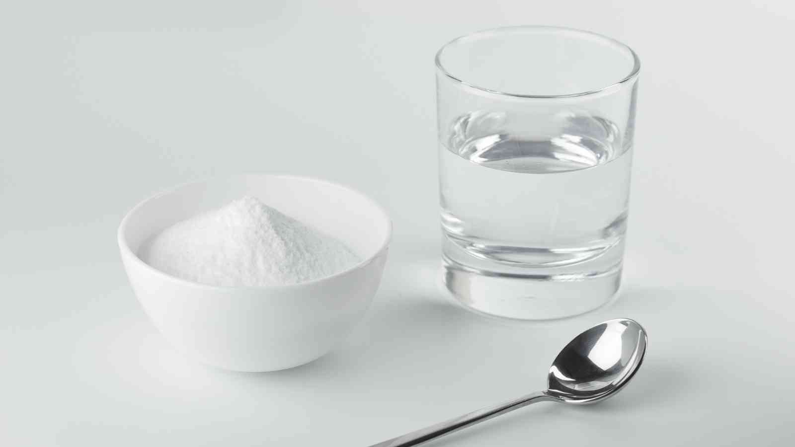 A bowl of baking soda, a glass water, and a silver spoon on a white surface.
