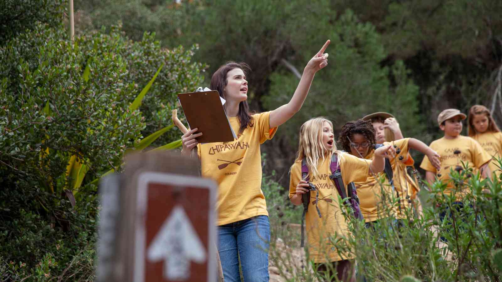 A group of children and a young woman in yellow shirts hike in a wooded area. The woman points ahead while holding a clipboard.