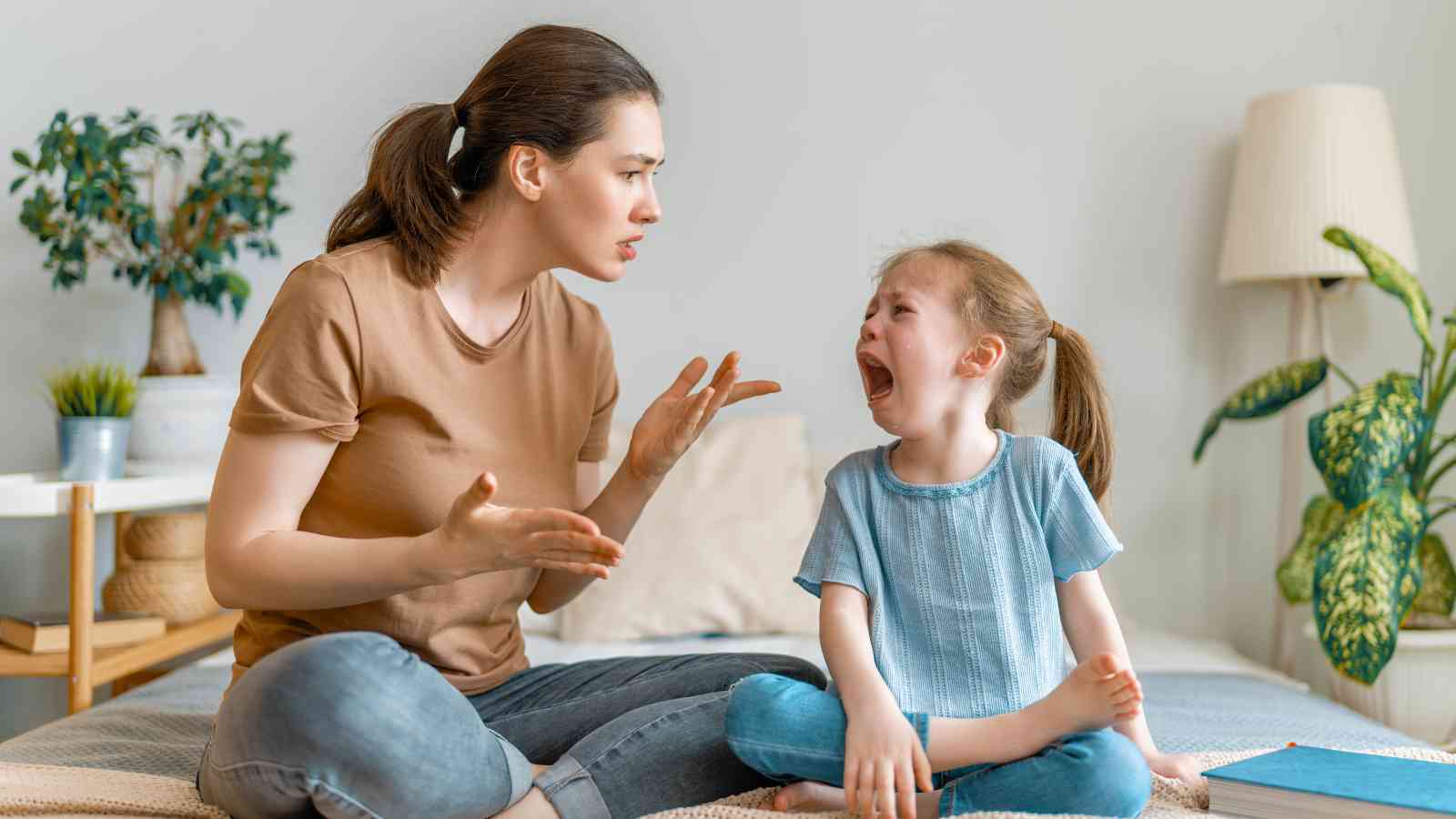 A woman is sitting on a bed, appearing angry and talking to a crying young girl seated next to her. The room has plants and a lamp in the background.