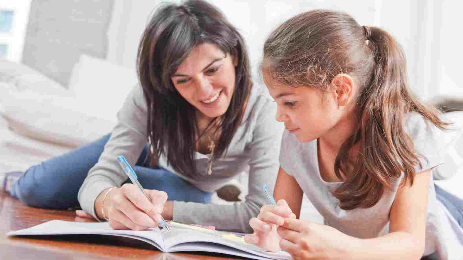A woman and a young girl are lying on the floor, writing in a notebook together.