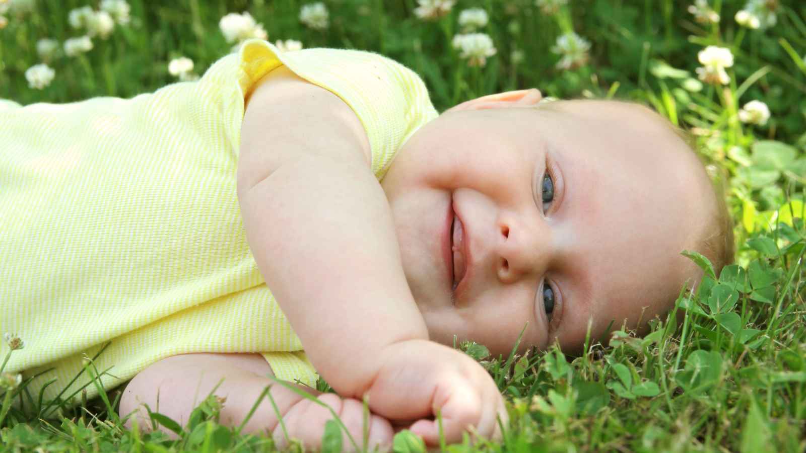 A baby in a yellow outfit lies on green grass, smiling.