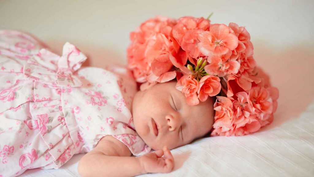 A baby sleeps on a white blanket wearing a pink floral dress, with a large wreath of pink flowers on her head.