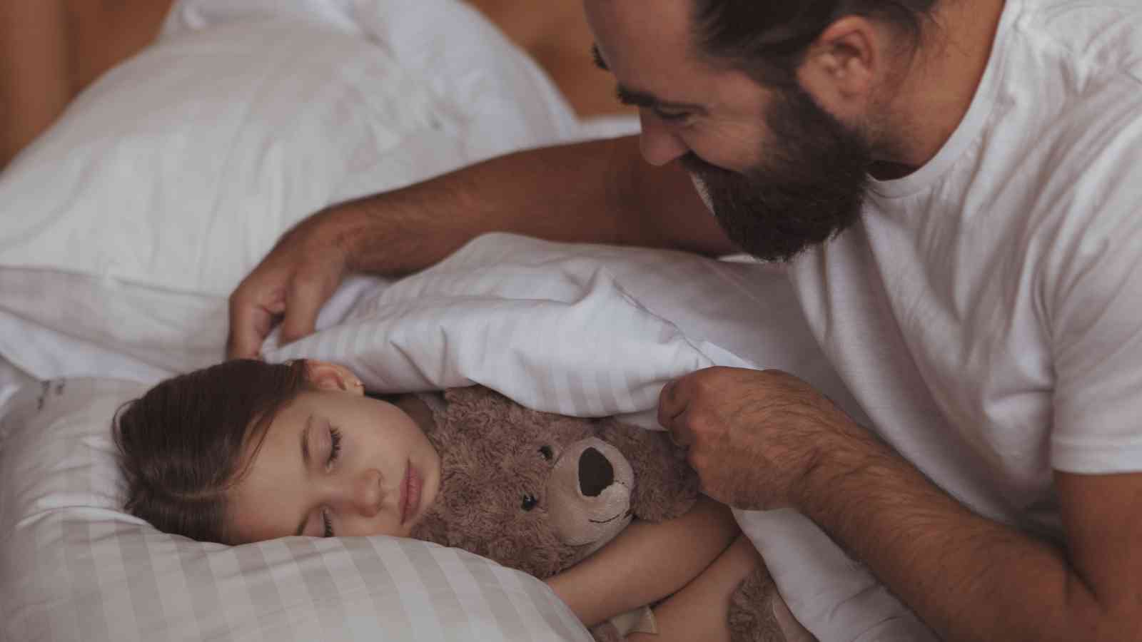A dad tucks his daughter holding a teddy bear into bed while she sleeps.