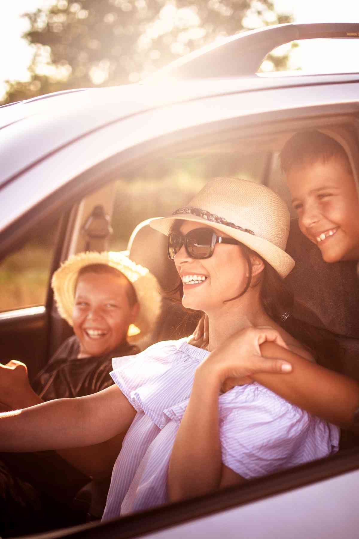 A woman wearing sunglasses and a hat is sitting in the driver's seat of a car, smiling, with two children in hats beside her, laughing.