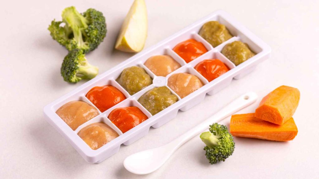 An ice cube tray filled with various purees, with carrots, broccoli, and a pear slice around it.