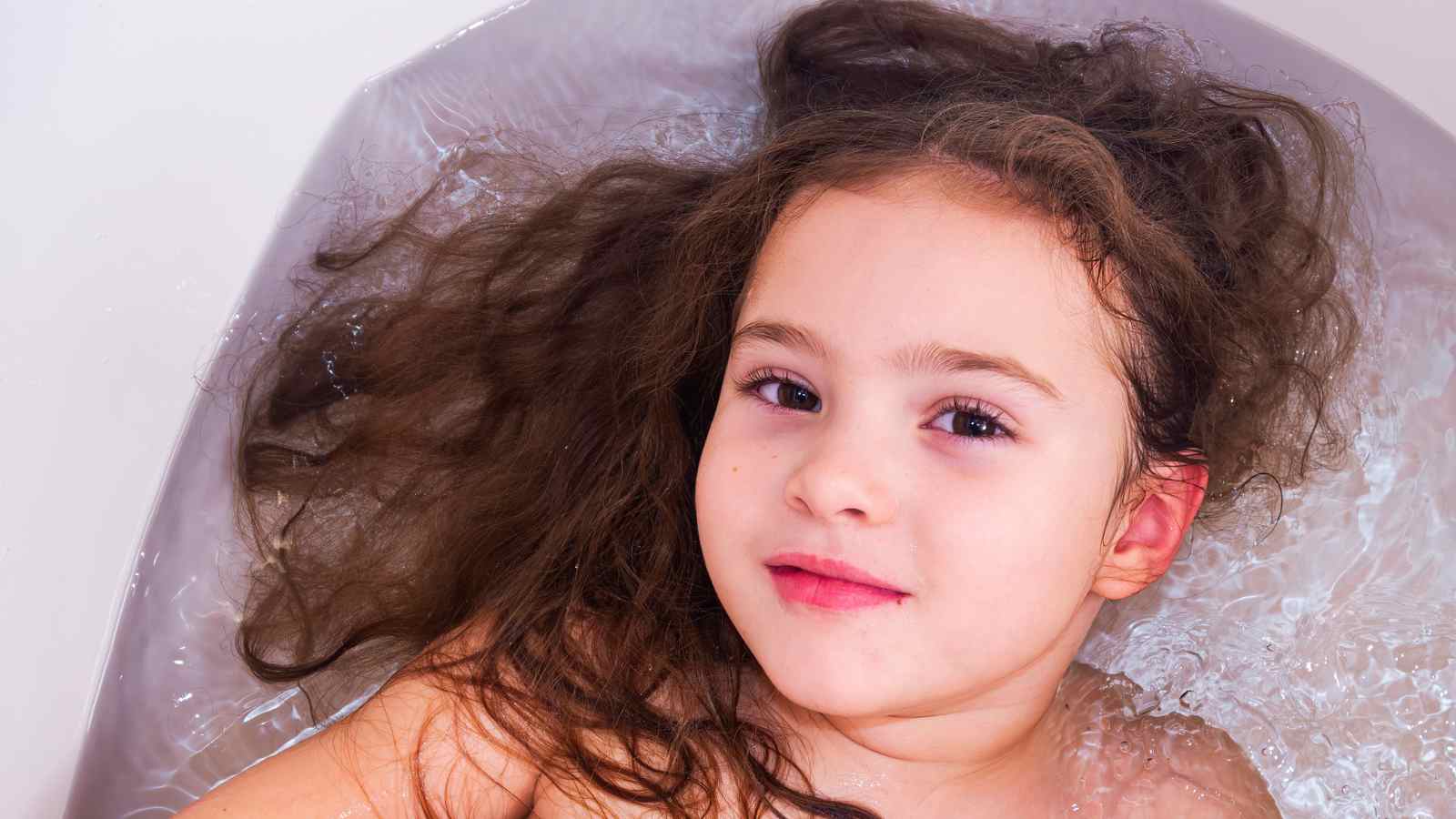 A young child with long, wet hair lies in a bathtub filled with water, facing the camera with a slight smile, enjoying the benefits of a detox bath for kids.