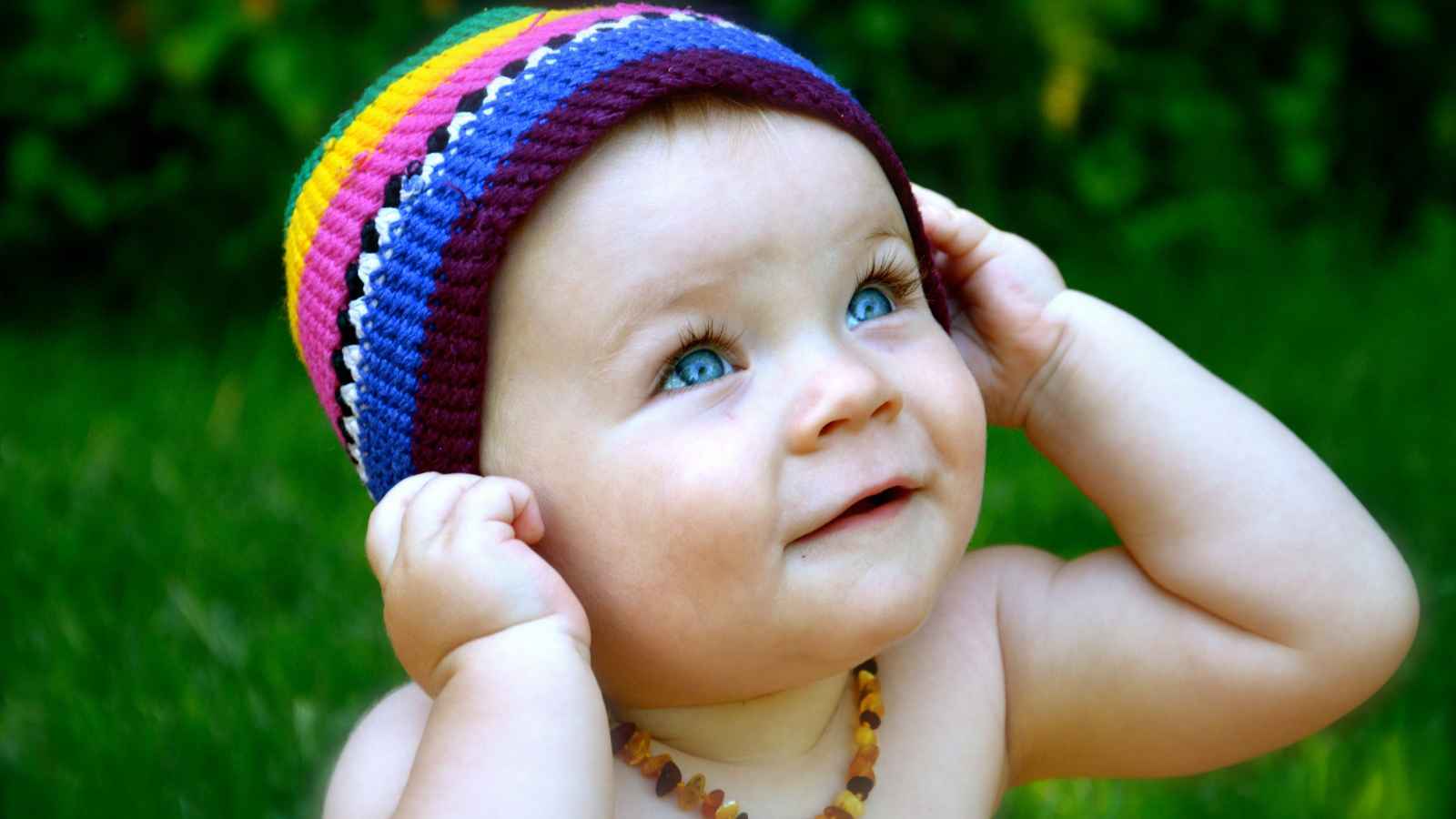 A baby with blue eyes wears a colorful knit hat and a necklace while looking up.