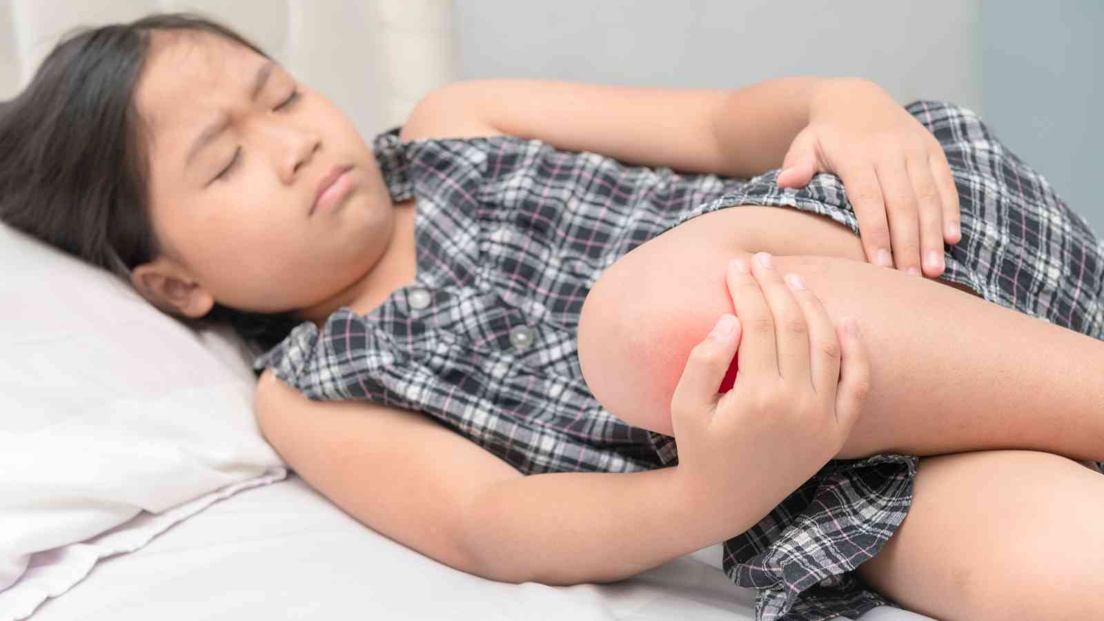 A child lies on a bed holding their red and swollen knee, appearing to be in discomfort.